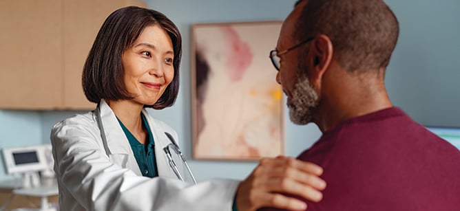 A physician smiling while placing a hand on a patient’s shoulder in an exam room